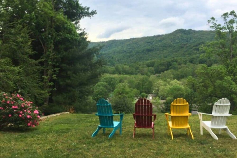 Four colorful Adirondack chairs are lined up on a green lawn overlooking a serene mountainous landscape.