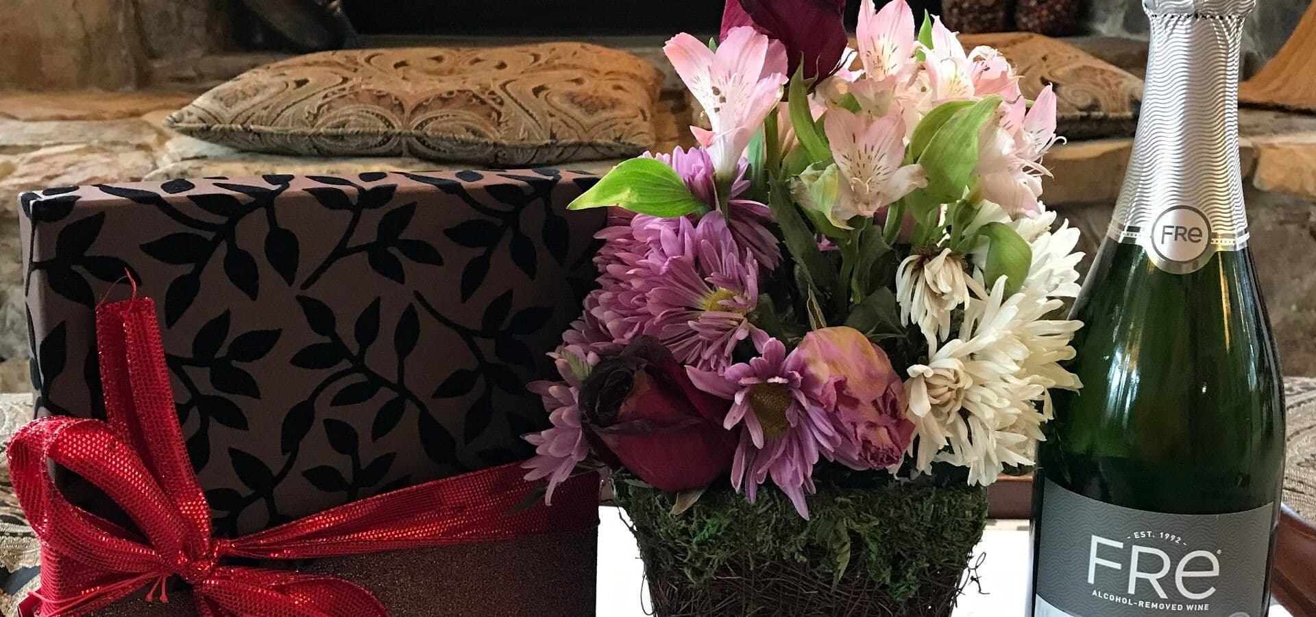 A decorative gift wrapped in black and red sits next to a floral arrangement and a bottle of non-alcoholic sparkling wine.