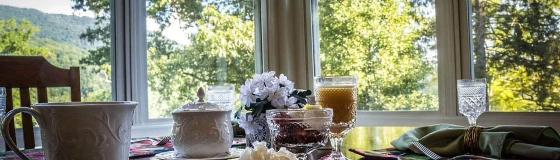 A table set for breakfast, featuring a cup, sugar bowl, pastries, and juice, with large windows overlooking greenery.