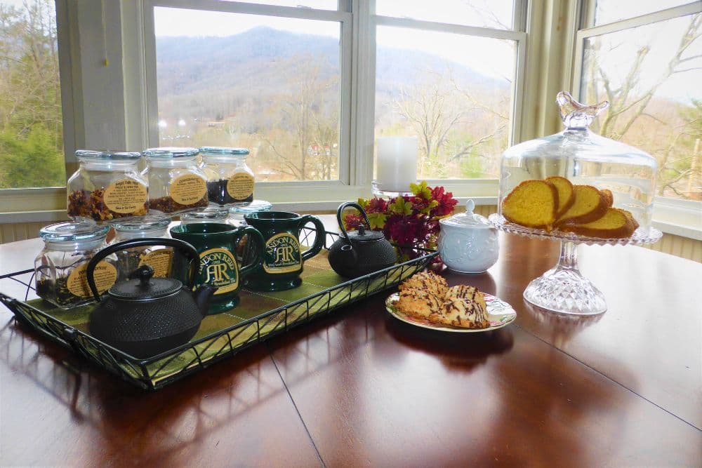 A cozy table setting featuring teapots, mugs, cookies, and a glass cake stand with sliced cake, overlooking a scenic view.