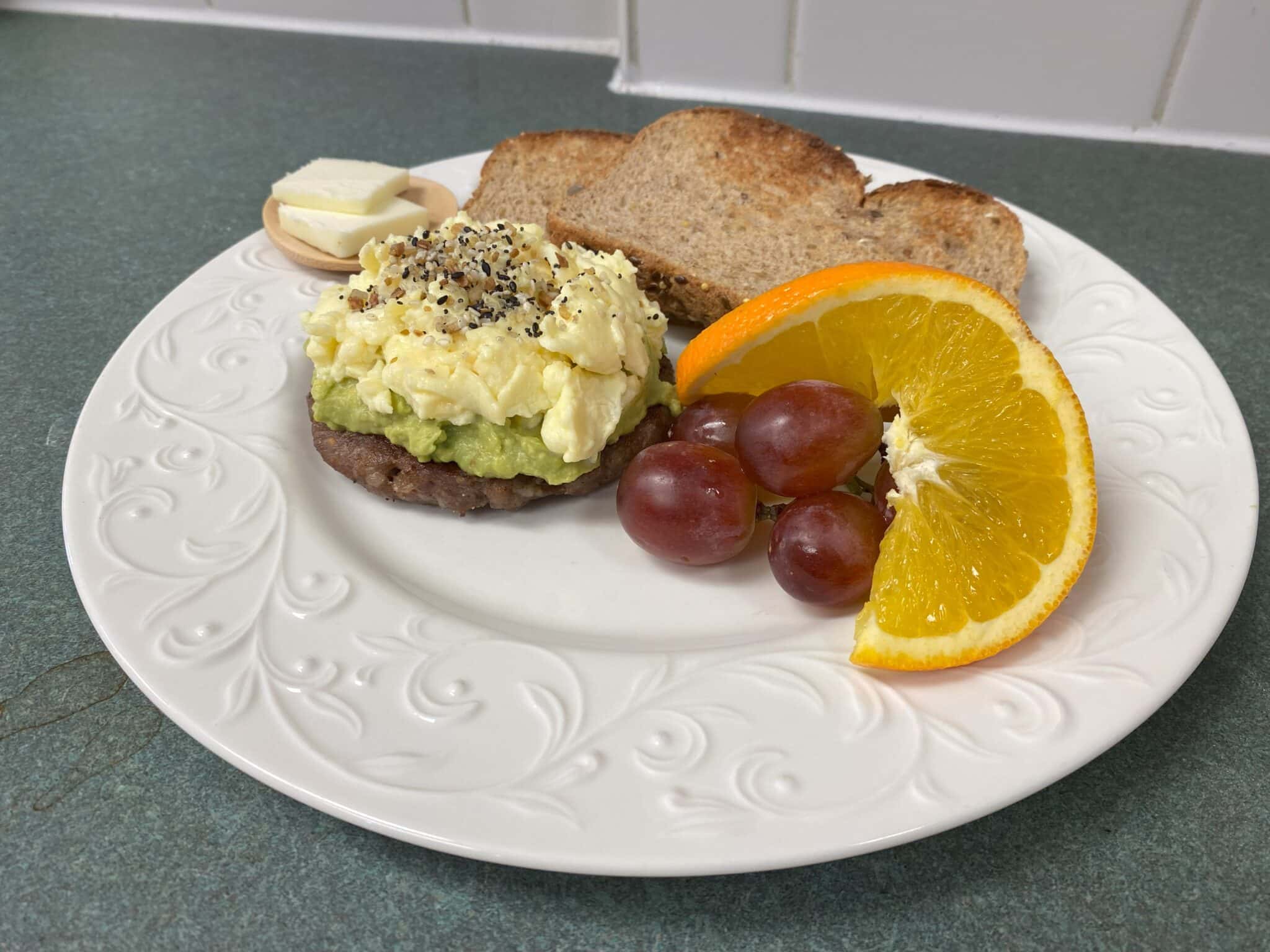 A plate with toasted bread, avocado and egg spread, served with grapes, an orange slice, and a small dish of butter.