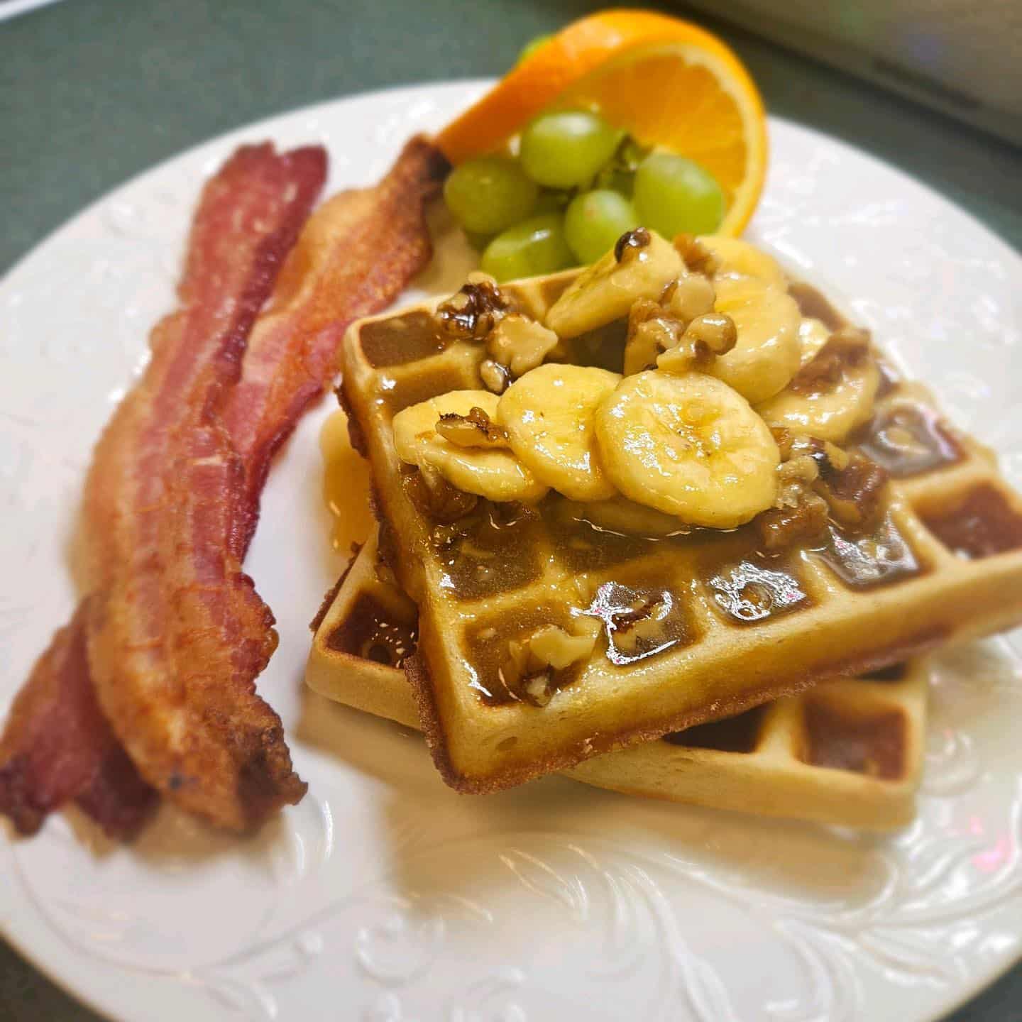 A plate featuring waffles topped with bananas and walnuts, accompanied by crispy bacon, grapes, and an orange slice.
