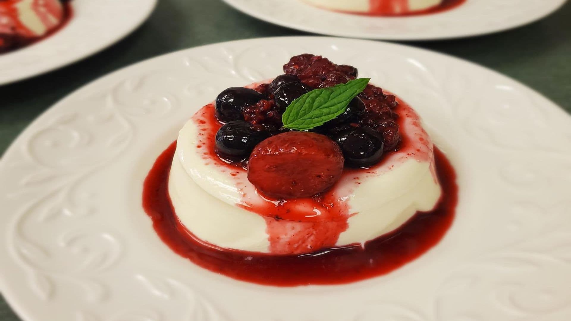 A creamy dessert topped with mixed berries and a mint leaf on a decorative plate.