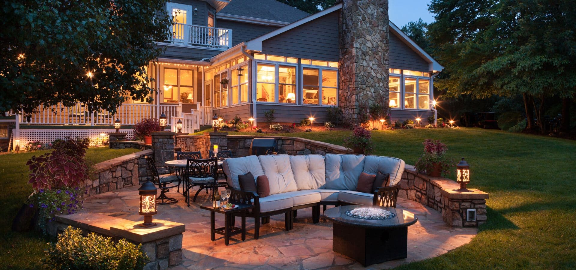 A cozy outdoor seating area with a fire pit, surrounded by lush greenery and illuminated by soft lighting at dusk.