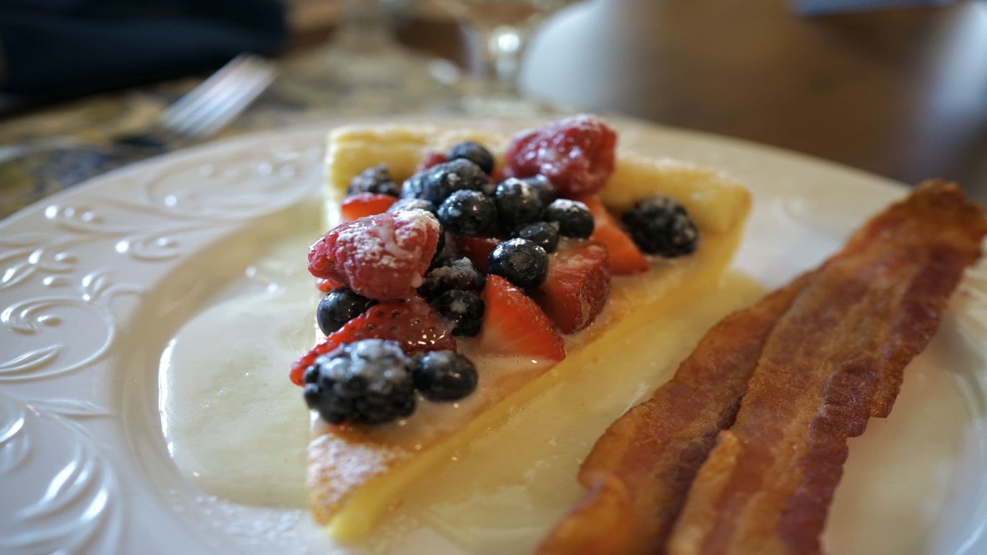 A slice of fruit-topped pastry with berries and a side of crispy bacon on a decorative plate.