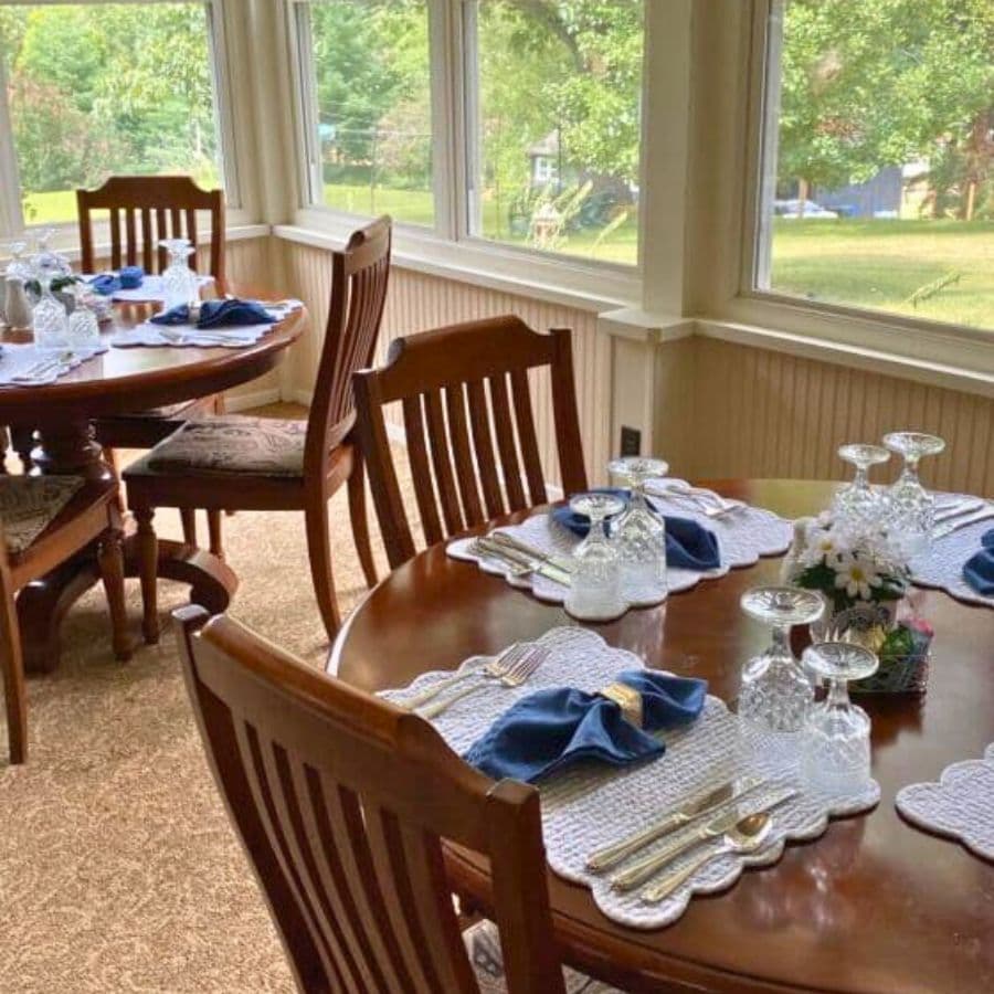 A cozy dining area with two wooden tables set elegantly, featuring blue napkins and glassware, surrounded by large windows overlooking greenery.