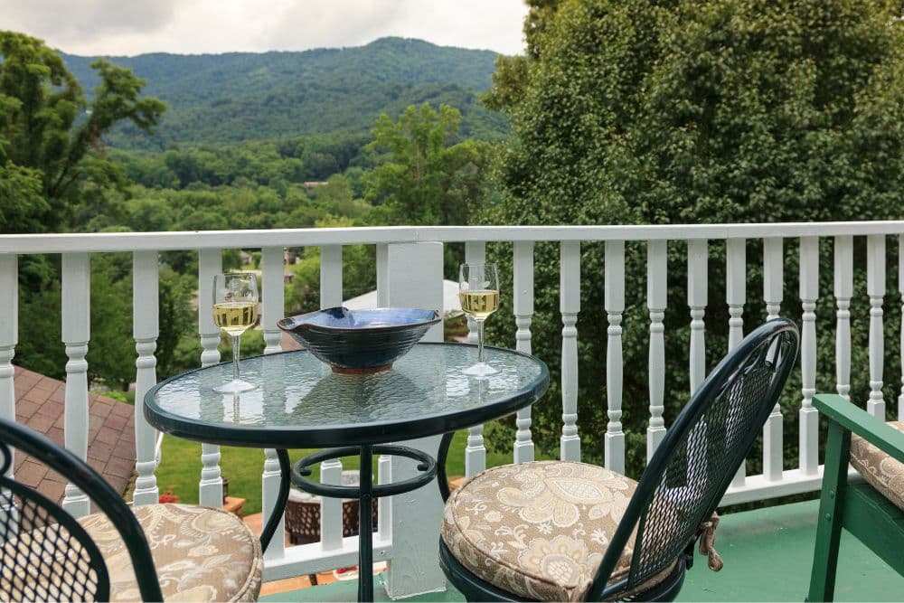 A small table with two glasses of wine overlooks a lush mountain landscape from a balcony.