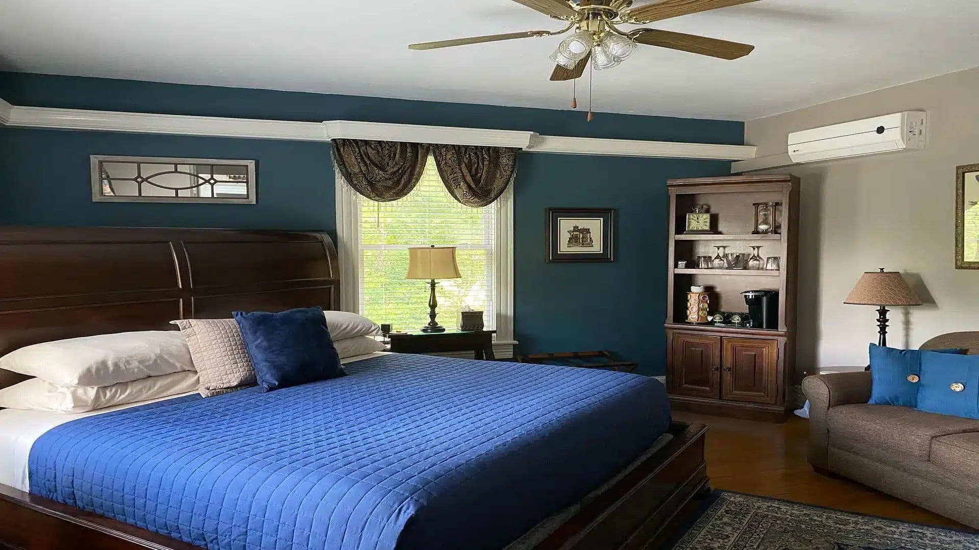 A neatly arranged bedroom featuring a large bed with a blue quilt, a wooden dresser, and a cozy seating area.